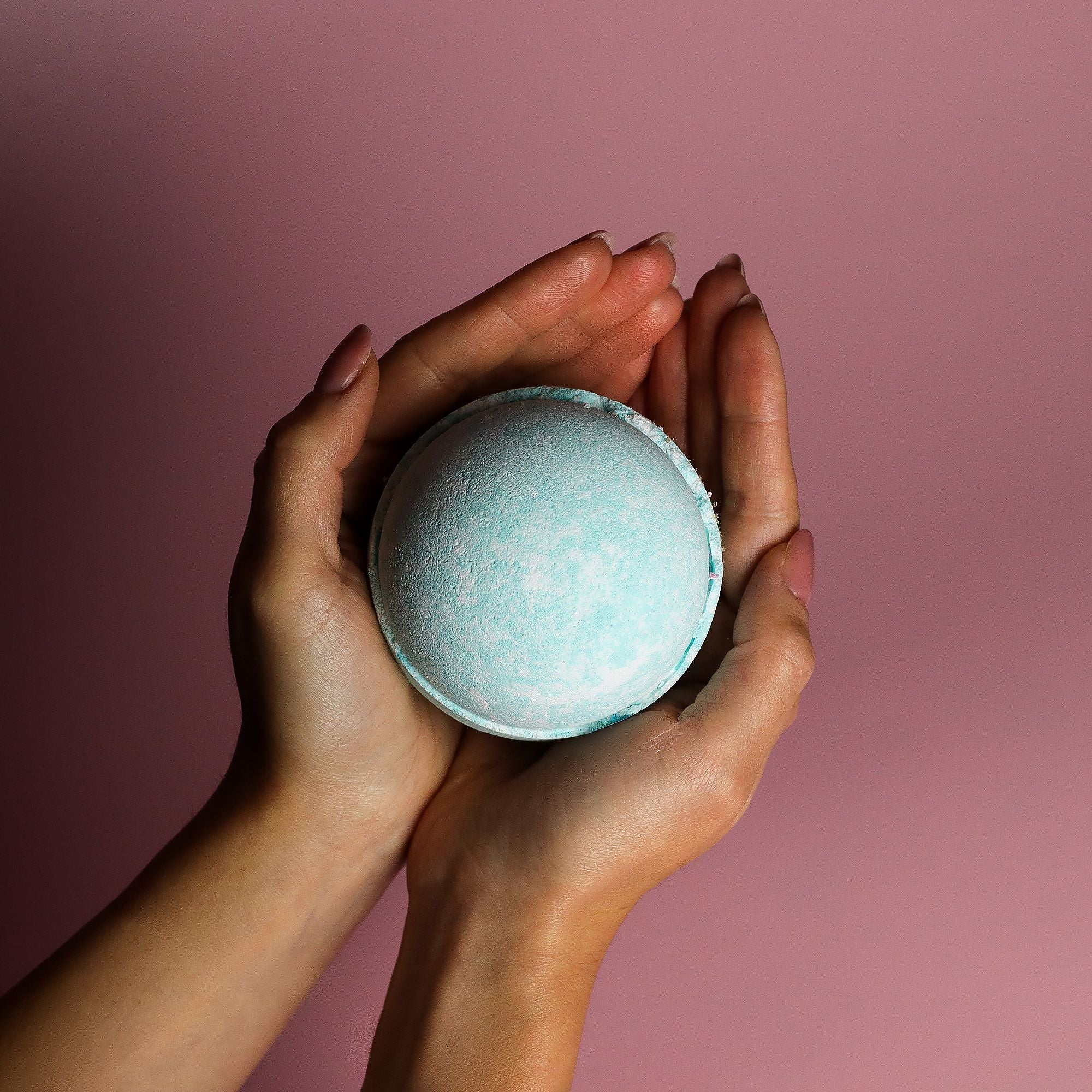 Hands holding a blue bath bomb against a pink background